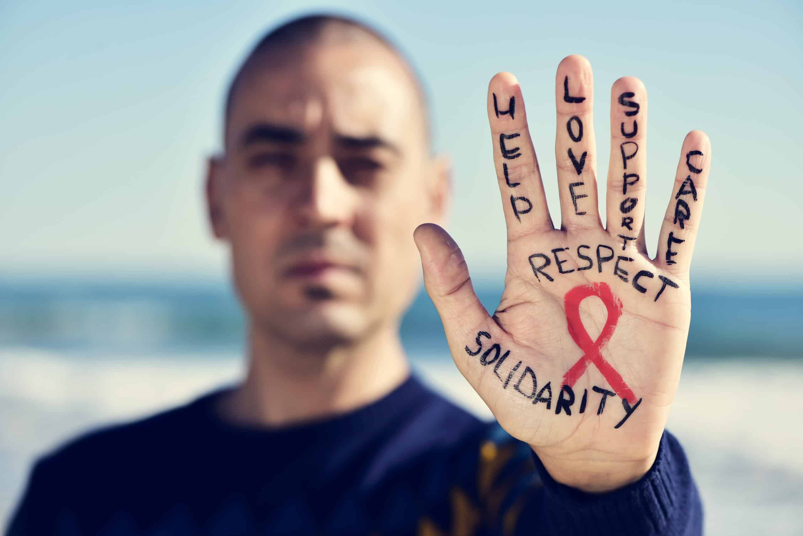 Primer plano de la mano de un joven caucásico en la que se ha pintado un lazo rojo en señal de lucha contra el sida, junto con las palabras «ayuda», «amor», «apoyo», «cuidado», «respeto» y «solidaridad».